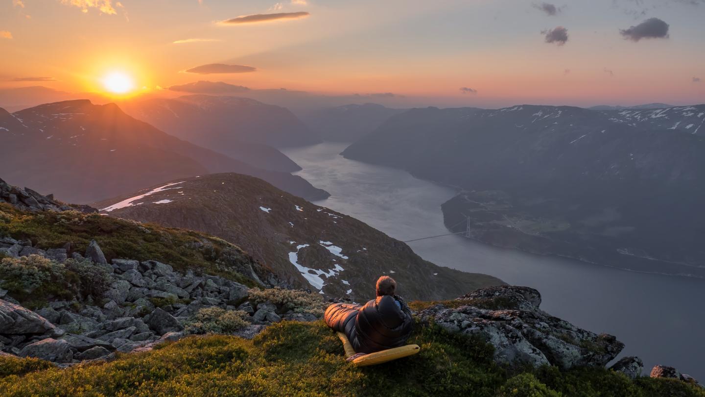 mann i sovepose på en fjelltopp som ser på solnedgangen over sørfjorden i Hardanger