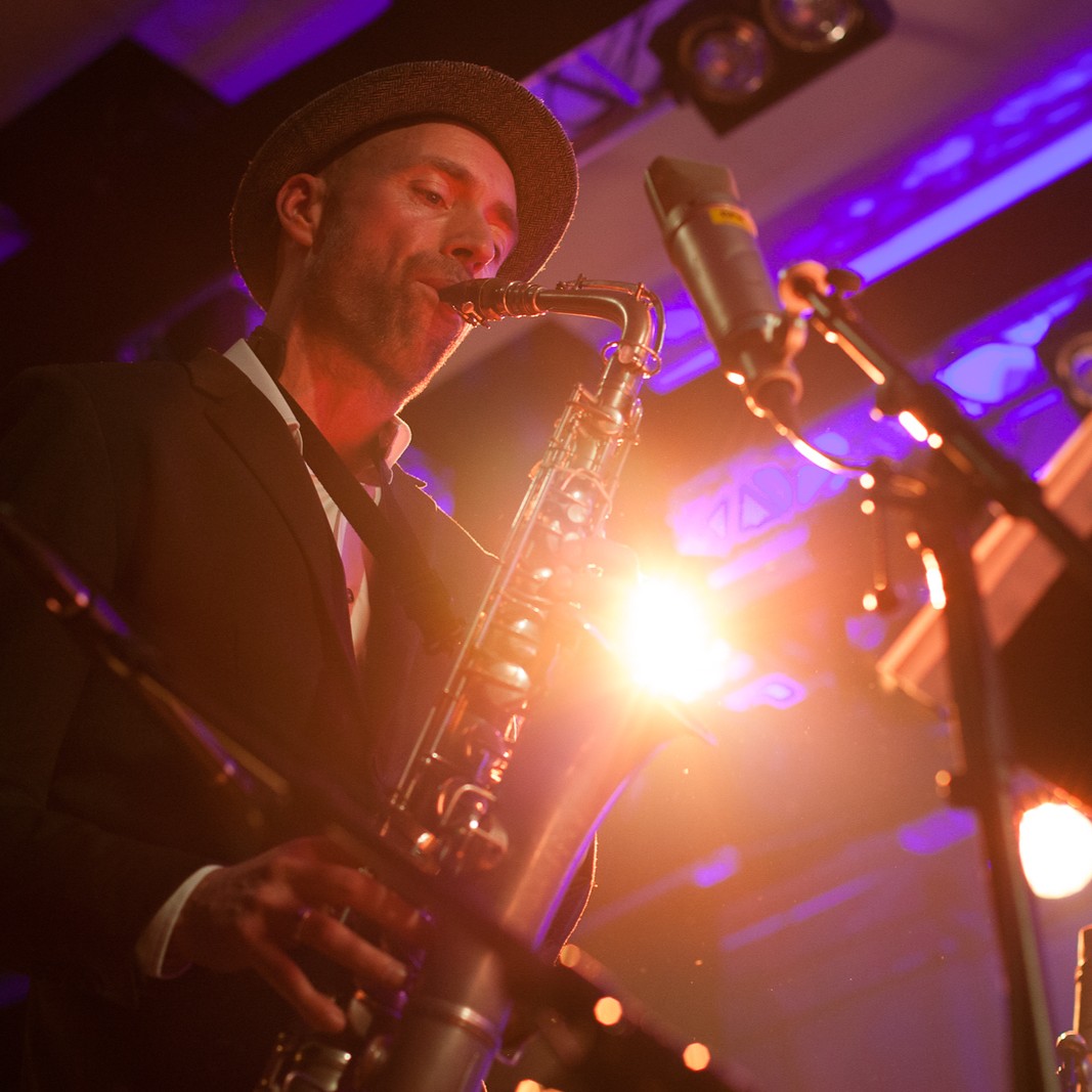 nærbilde av en mann, med hatt, spiller trompet. I bakgrunnen ser man scenelysene.