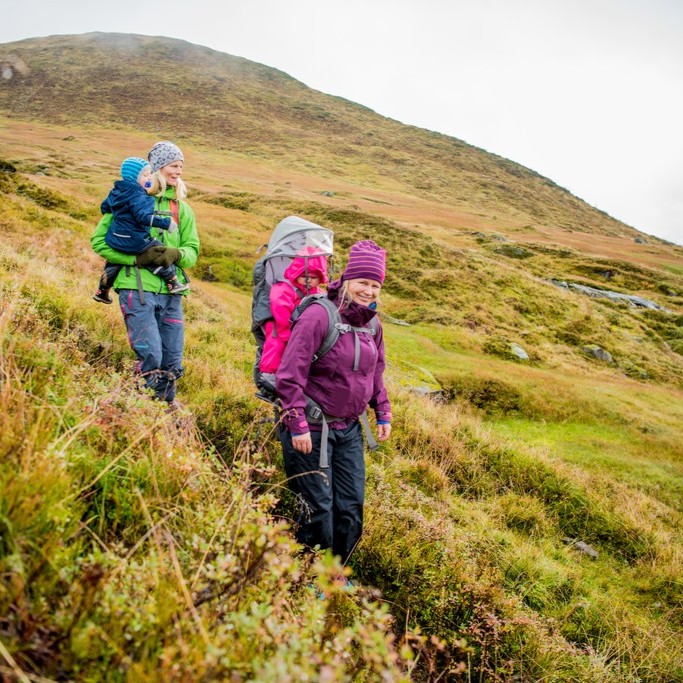 To damer med kvar sitt barn, på fjelltur i haustvær. Fargane i landskapet er litt meir haustlege enn på sommaren og dei har på seg allværsklede.