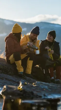tre jenter tar en pause på turen. De har fargerike klær, ler og nyter utsikten. I bakgrunnen ser man Lønahorgi