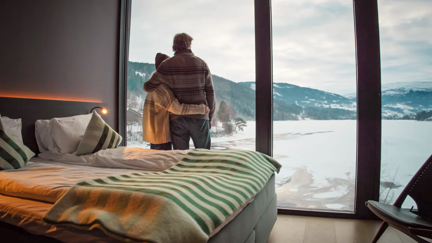 par nyter utsikten fra rommet sitt på elva hotell. I forgrunnen ser man sengen plassert like ved de store vinduene og utenfor et nydelig isdekt vann, en ås og noen snøkledde fjell