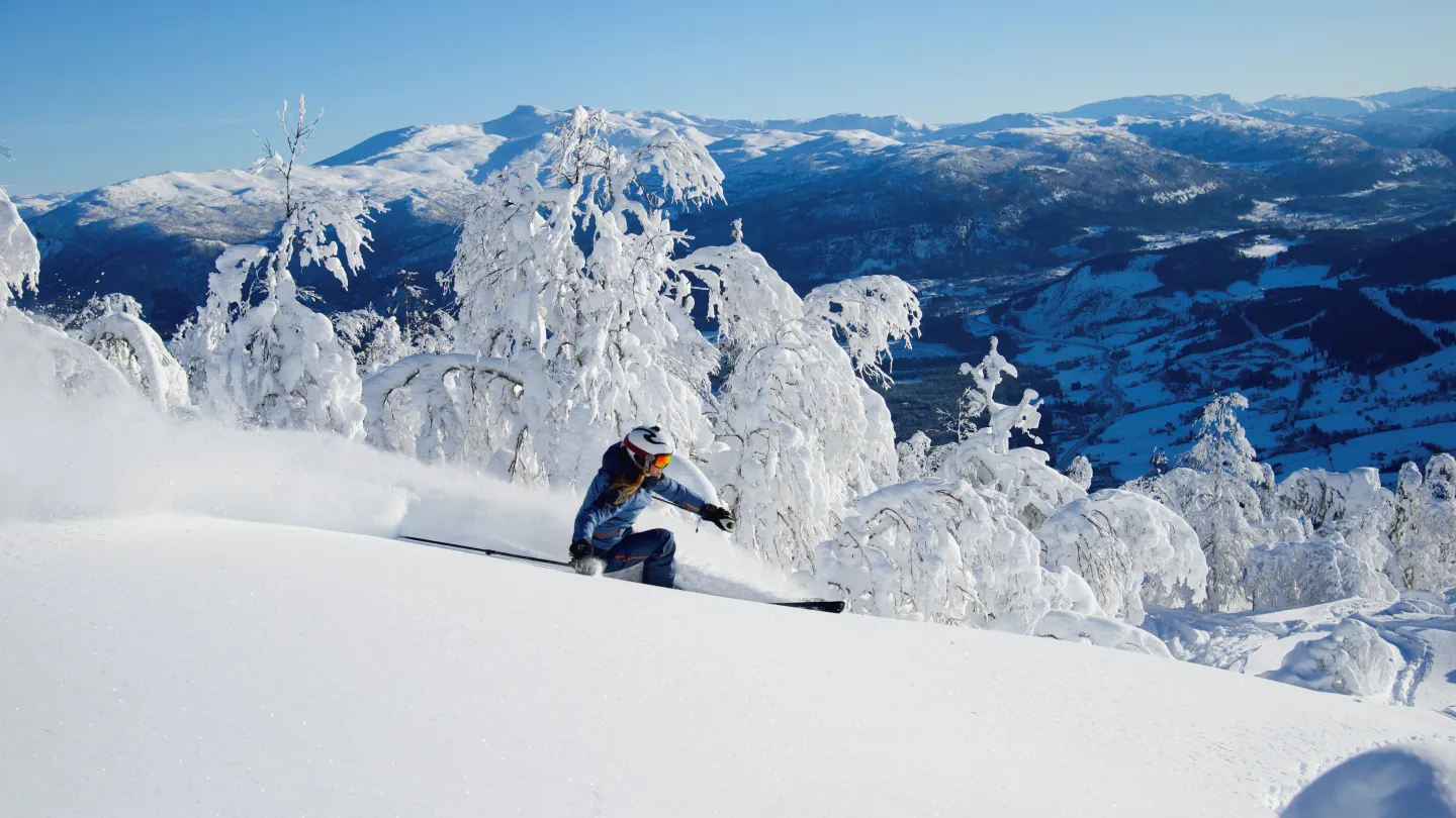 kvinne opp piste i voss resort, snøen står opp bak henne og solen skinner
