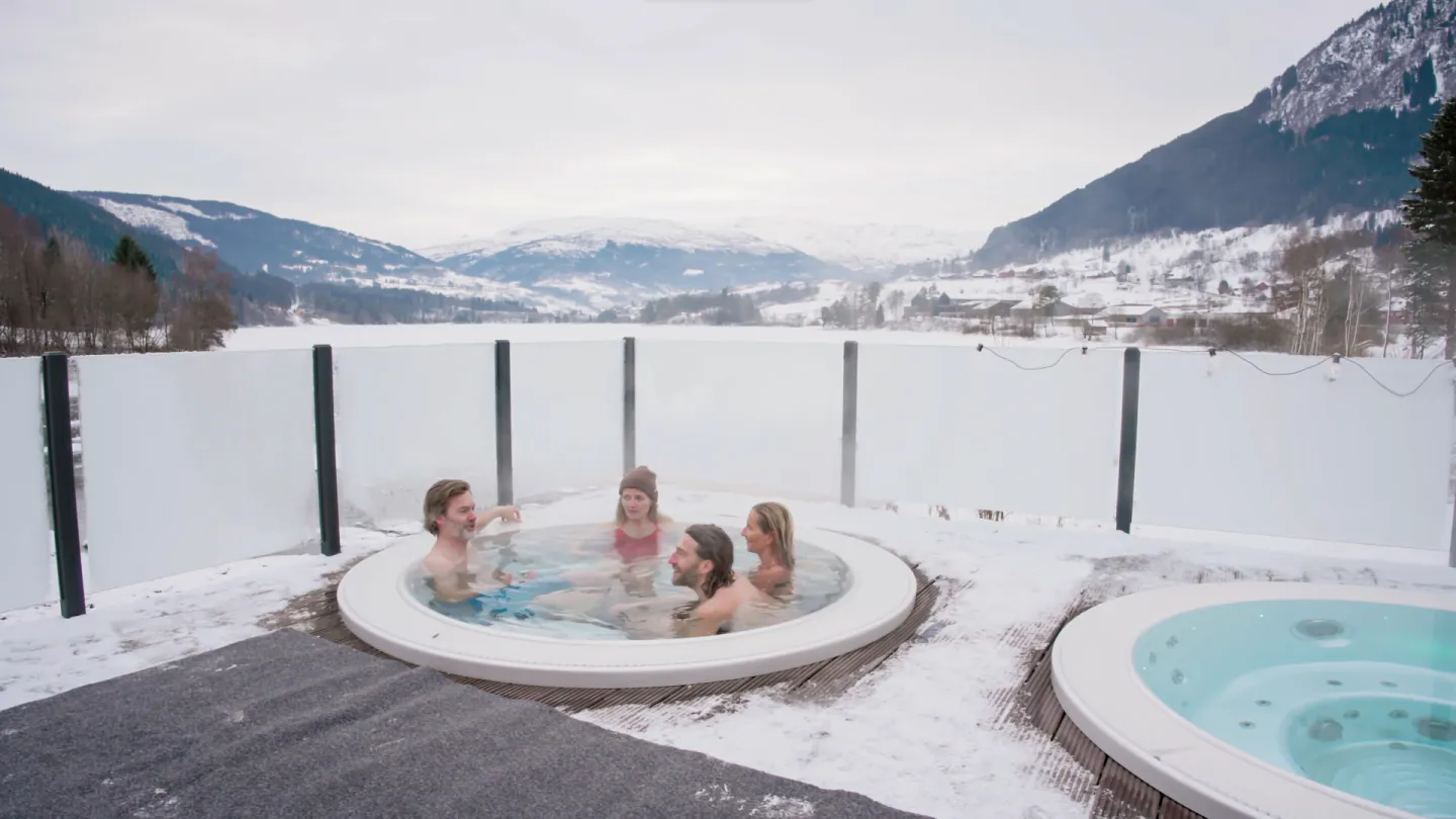 Fire voksne i utendørs boblebad på Elva Hotel, med flott utsikt utover vatnet og fjella. 