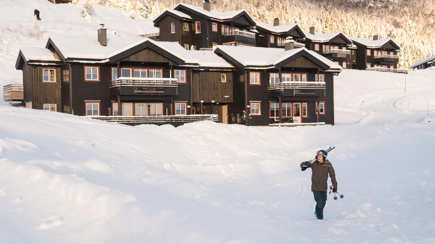 mann går fra hytte, med skiene på skulderen, klar for skibakken. 