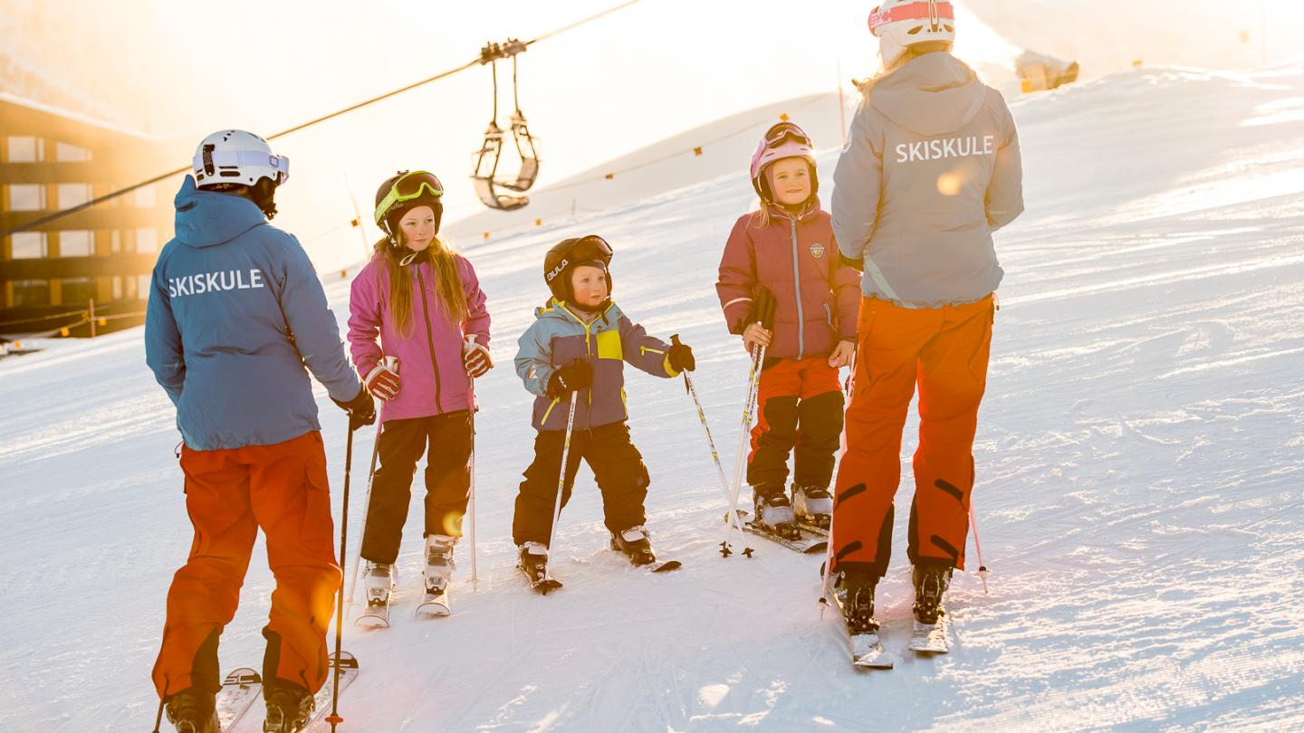 Tre barn står i skibakken og får opplæring av to jenter frå skiskulen.