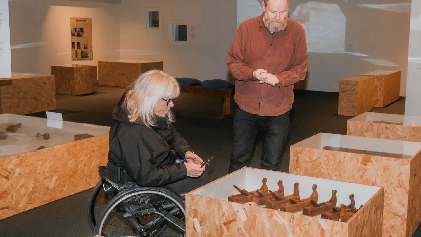Dame i rullestol og gående mann ser på utstillingen hos Voss Folkemuseum