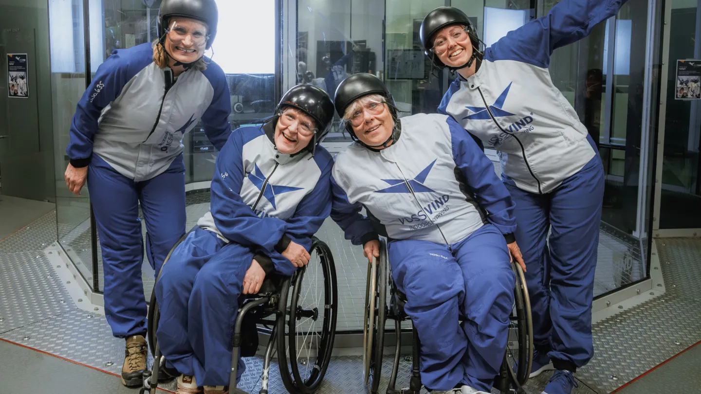 fire damer står forann vindtunnelen, klare for å fly, de to i midten sitter i rullestol. 