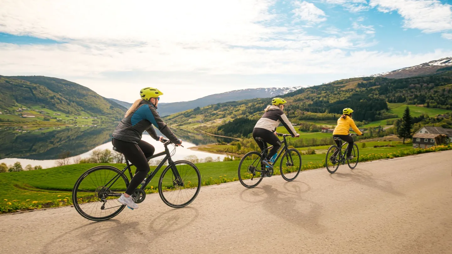 Tre damer sykler langs veien på Voss. I bakgrunnen ser mann Vangsvatnet og fjellene. 